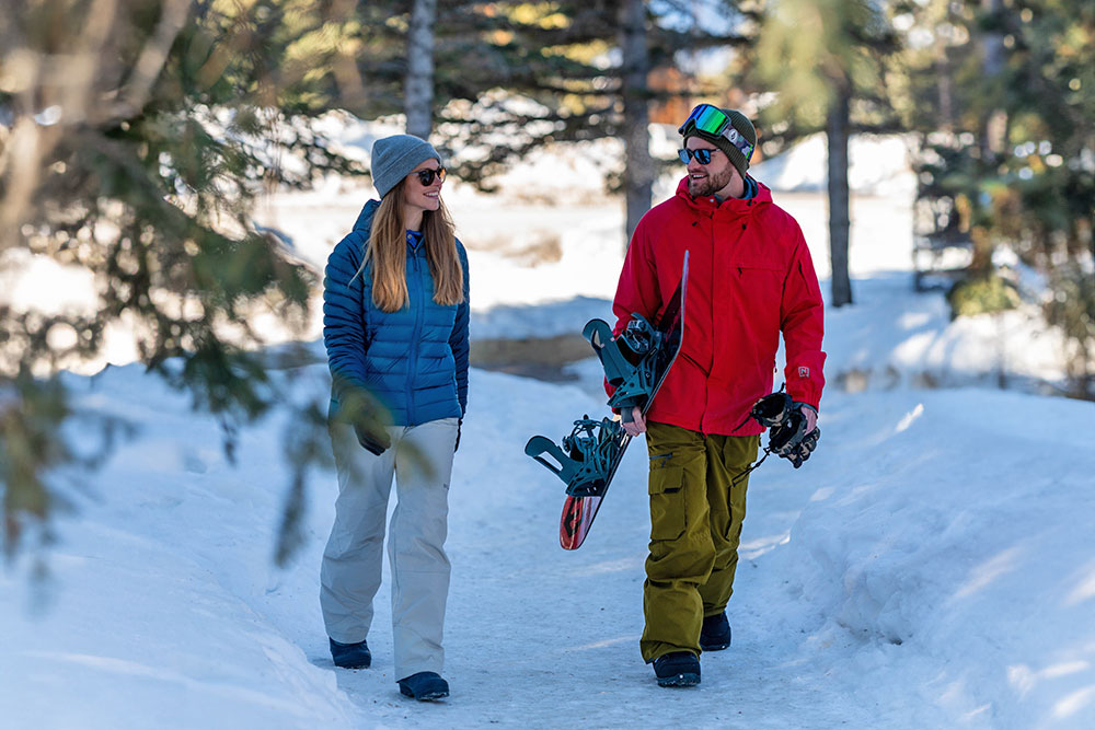 2 people walking on snow path surrounded by trees in winter, one person is holding a snowbaord